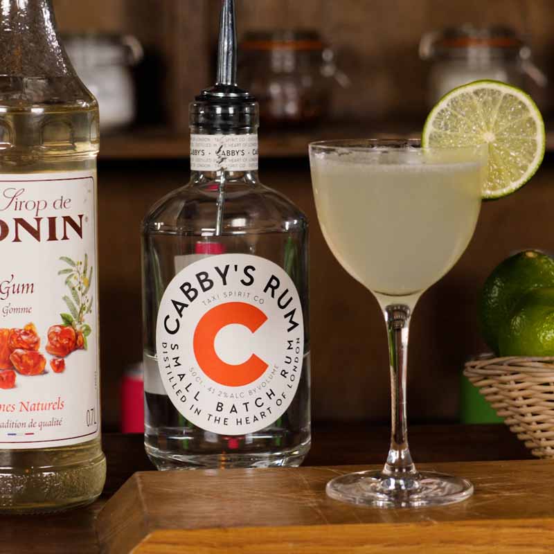 Daiquiri cocktail on a bar top with a bottle of Cabby's Rum and some limes