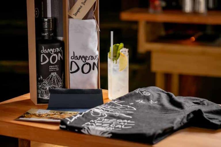 A bottle of Dangerous Don Mezcal on a table with a cocktail and a t-shirt